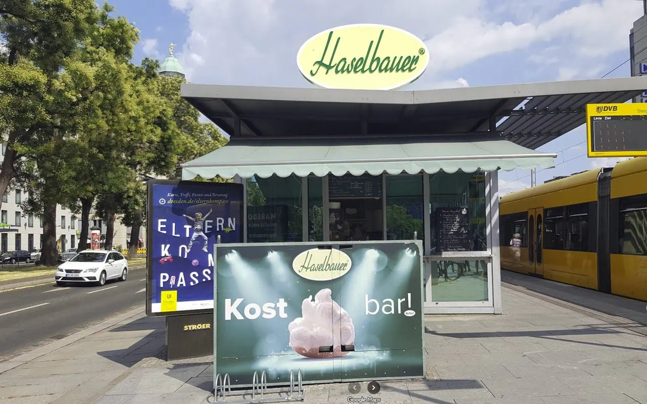 Eis-Pavillon Haselbauer Eiscreme-Stand mit grünem Vordach und gelbem Logo, Frontansicht am Straßenbahn-Bahnhof, Werbung und Umgebung sichtbar