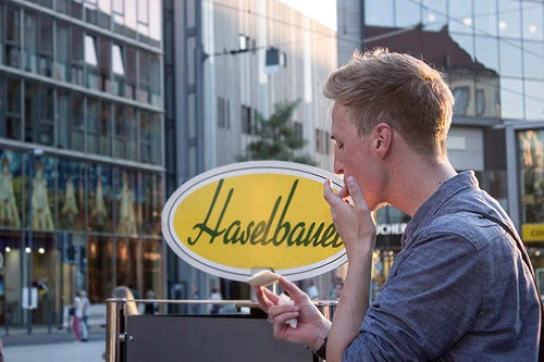 Haselbauer Eis Logo Schild in Gelb-Weiß vor moderner Glasfassade in einer urbanen Straßenszene; junger Mann im Vordergrund deutet auf das Schild und schaut darauf